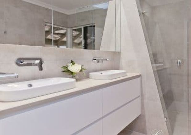 Contemporary bathroom with a long white floating vanity, two above-counter basins, wall-mounted chrome taps, a large mirrored wall, and a glass-enclosed shower. Pale grey-beige tiled walls and a light neutral palette create a clean, refined appearance.