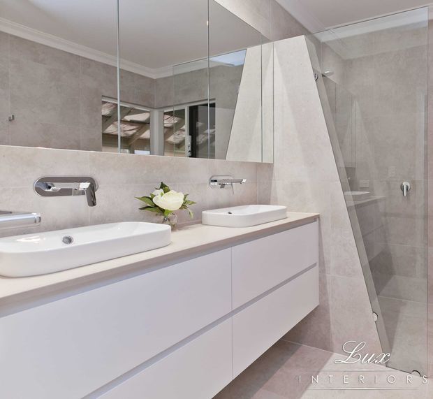 Bright contemporary bathroom featuring a double floating vanity with white vessel basins, wall-mounted chrome tapware, large mirrored cabinetry, and a frameless glass shower enclosure. The room is finished in large-format light beige-grey stone-look tiles with a crisp, refined appearance.