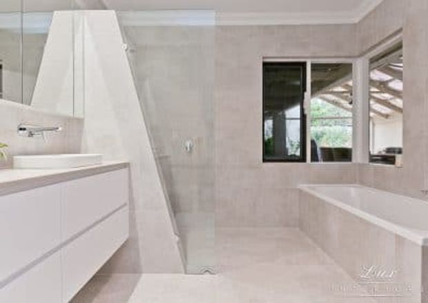 Bright contemporary bathroom with a freestanding white bathtub, floating white vanity, pale stone or large-format tiled walls and floor, frameless shower glazing, and black-framed windows overlooking a covered outdoor area.