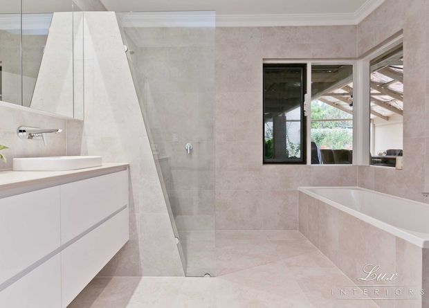 Bright contemporary bathroom with pale stone-look wall and floor finishes, a frameless glass shower, freestanding white basin on a floating vanity, wall-mounted tapware, and a built-in bathtub beneath a window. A large mirror and reflected glazing add spaciousness, while the neutral palette creates a refined, clean atmosphere.