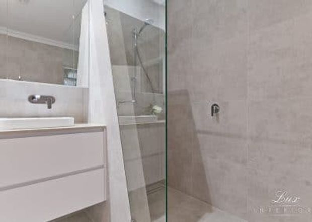 Compact contemporary bathroom featuring a wall-mounted white vanity with integrated basin and chrome tapware, a frameless glass shower enclosure, and pale beige stone-look wall tiling. A large mirror reflects the vanity and shower area.