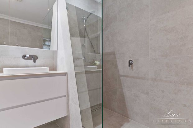 Contemporary bathroom featuring a floating pale vanity with integrated basin, wall-mounted chrome tapware, large-format light stone-look wall and floor tiles, and a frameless glass shower enclosure. A mirrored wall adds depth to the compact, refined space.