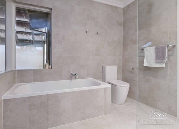 A bright contemporary bathroom with full-height beige stone-look wall and floor tiles, a built-in bathtub, wall-mounted bath fittings, a compact toilet, and a frameless glass shower enclosure. A towel rail with folded towels is mounted inside the shower area, and a window provides natural light.