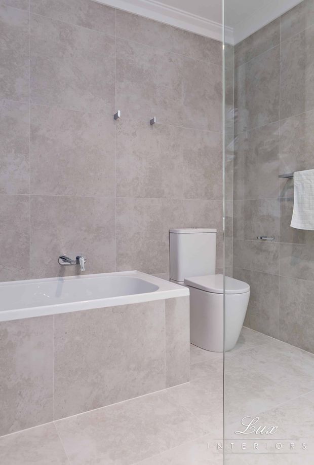 A bright, contemporary bathroom featuring large-format beige stone-look tiles on the walls and floor, a white built-in bathtub, a modern white toilet, chrome tapware, and a frameless glass shower screen. The space has clean lines and a neutral, polished finish.