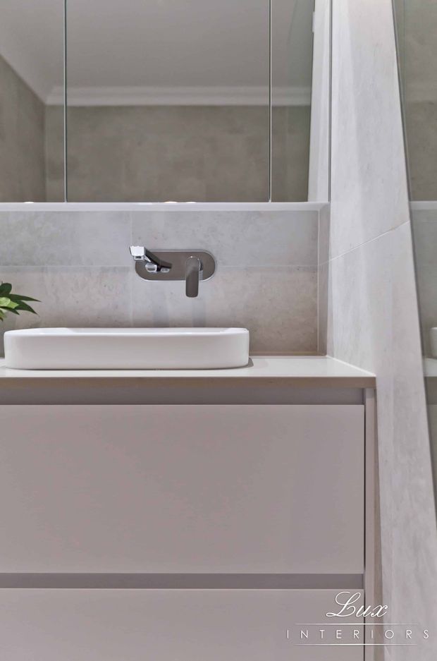 A compact contemporary bathroom vanity featuring a white vessel basin on a pale stone benchtop, wall-mounted brushed metal tapware, large mirrored cabinetry, and soft beige stone-look wall tiling. The vanity has minimalist flat-front cabinetry in a pale neutral finish, with a small plant adding a natural accent.