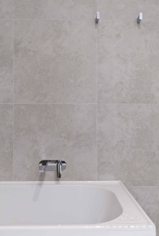 Close-up of a white bathtub against a wall clad in large-format pale beige-grey stone-look tiles. A wall-mounted chrome bath mixer and spout are visible, along with two small chrome wall hooks. The image has a clean, understated bathroom finish with minimal decoration.