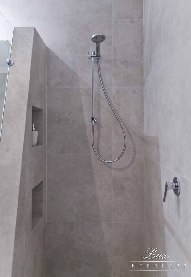 A compact modern shower enclosure clad in large-format pale grey-beige stone-look tiles. The shower features a chrome handheld shower on a vertical rail, recessed wall niches with toiletries, a chrome control, and a partial glass screen. The space is clean, restrained, and neutral in appearance.