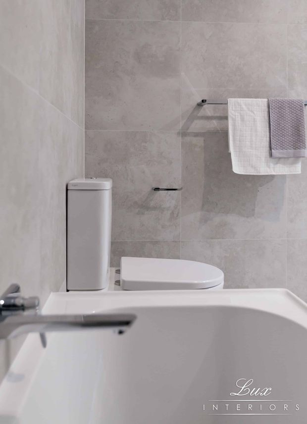 Compact modern bathroom featuring a white bathtub in the foreground, a close-coupled white toilet, pale grey-beige large-format tiled walls, and minimalist chrome towel hardware with folded towels. The space has a clean, bright, understated appearance.