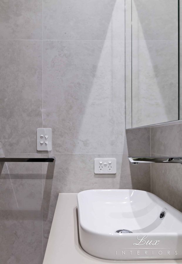 Close-up of a compact contemporary bathroom vanity area with a white ceramic vessel basin on a pale benchtop, grey-beige tiled walls, a mirrored cabinet, chrome tapware, and white electrical outlets. The image has a clean, neutral, softly lit finish.