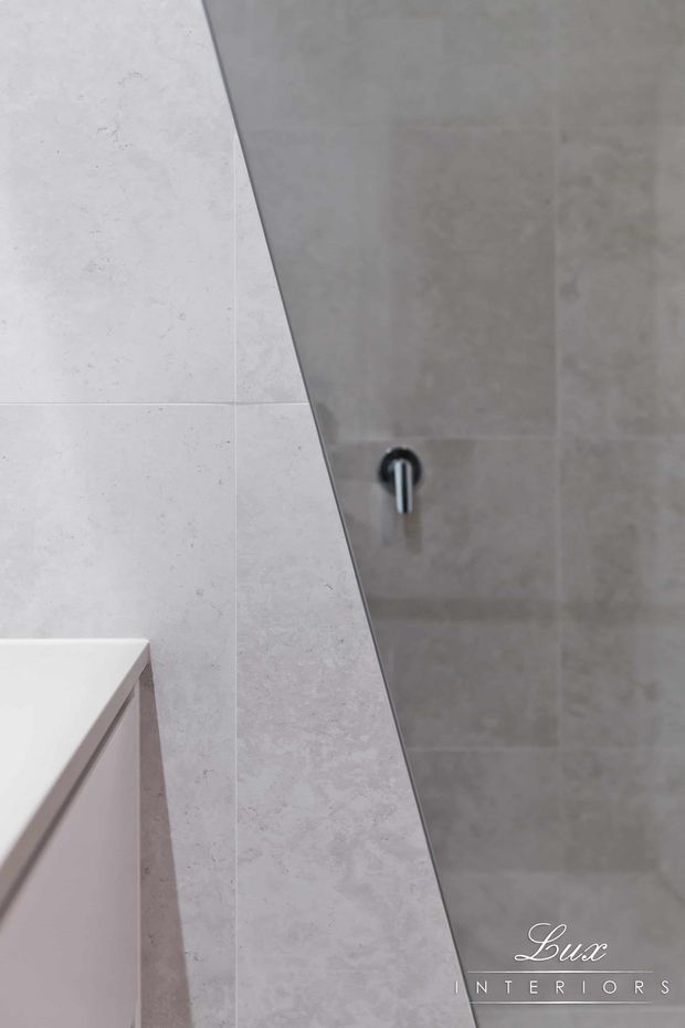 Close-up architectural interior detail showing a minimalist bathroom wall clad in large-format pale grey and warm grey tiles, with a sharp angled tiled partition or vanity edge and a dark metal wall control. A small portion of white built-in cabinetry or vanity is visible at the lower left. The image has a refined, understated contemporary aesthetic.