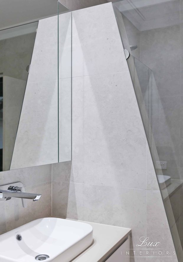 Close-up view of a contemporary bathroom featuring a white vessel basin on a light stone vanity, pale stone wall tiling, a frameless glass shower enclosure, large mirror panels, and minimalist chrome tapware. The angular tiled wall and clean neutral palette create a refined, modern interior.