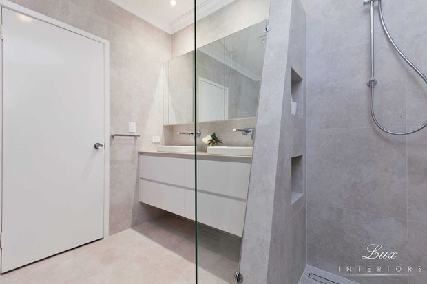 Bright contemporary bathroom with pale stone-look wall and floor finishes, a floating white vanity with integrated basin, mirrored wall cabinetry, a frameless glass shower screen, and recessed shelving built into a tiled partition. The room has clean, minimal detailing and soft neutral tones.