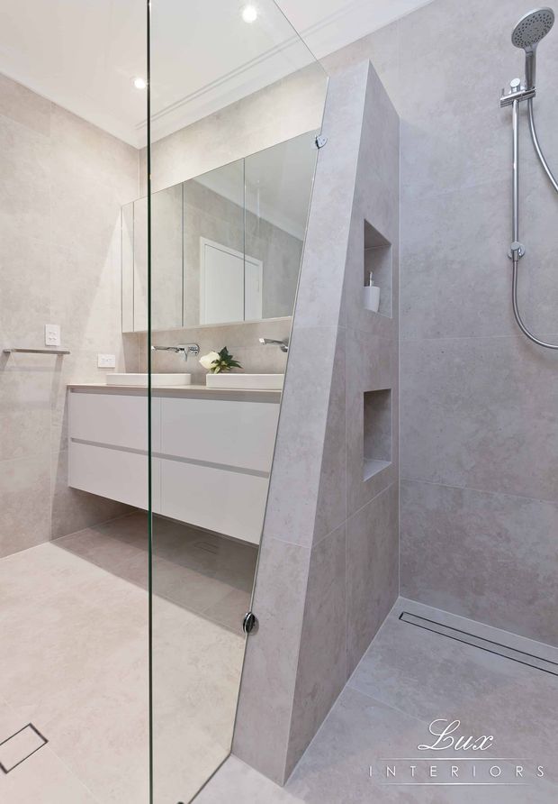 A bright, contemporary bathroom featuring a frameless glass walk-in shower, large-format pale stone-look wall and floor tiles, a wall-hung white vanity with integrated basins, mirrored wall cabinetry, recessed shower niches, and chrome fixtures. The layout uses a partial angled partition to separate the shower from the vanity.