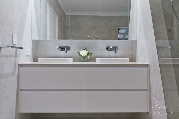 Contemporary double-vanity bathroom with a wall-mounted pale grey vanity, two white countertop basins, chrome wall-mounted taps, large mirrored cabinetry, and grey stone-look wall and floor finishes. A glazed shower enclosure is visible at right.