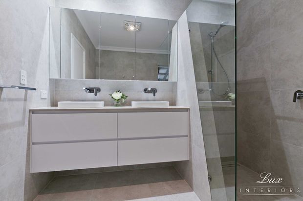 Contemporary double-vanity bathroom with a wall-mounted pale grey vanity, two white basins, mirrored cabinetry or wall panels above, and a frameless glass walk-in shower. Large grey stone-look tiles cover the walls and floor, with sleek chrome fixtures and soft integrated lighting.