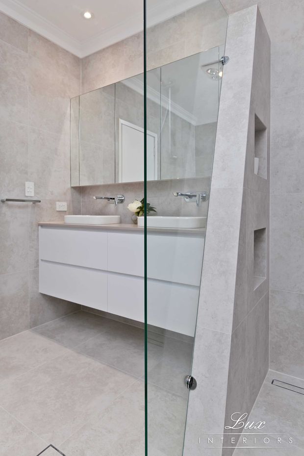 A bright contemporary bathroom featuring pale stone-look floor and wall tiles, a floating white vanity with integrated basin, mirrored wall cabinetry, wall-mounted chrome tapware, and a frameless glass shower screen. Built-in open niches provide shower storage, while recessed ceiling lighting and white cornices create a clean, refined finish.