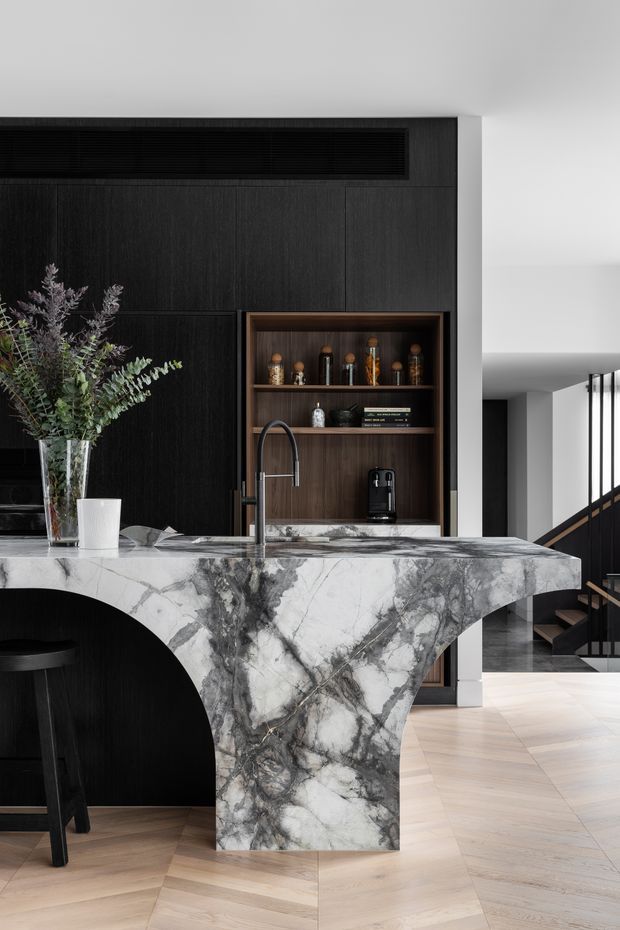 A sophisticated contemporary kitchen and living area featuring a dramatic veined stone waterfall island with a curved arch detail, dark timber wall cabinetry, an open drinks display niche, and a black gooseneck tap. Pale herringbone timber flooring contrasts with the dark joinery and stone. A stairway with black balustrading is visible in the background, while subtle greenery and countertop accessories add warmth.