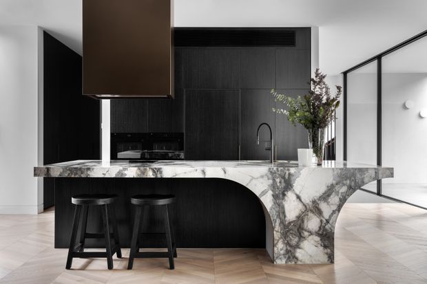Contemporary kitchen featuring a dramatic veined stone island with a sculptural curved base, dark timber cabinetry, black bar stools, and a dark suspended rangehood. The kitchen opens toward a bright adjoining space through black-framed glazing, with warm herringbone timber flooring and minimal monochromatic styling.