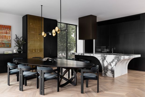 A sophisticated open-plan dining and kitchen interior. A dark sculptural dining table with upholstered chairs sits in the foreground, while black full-height cabinetry, a warm timber-toned display panel, marble-patterned island surfaces, and dramatic pendant lighting define the space. Large black-framed windows bring in natural light and greenery views.