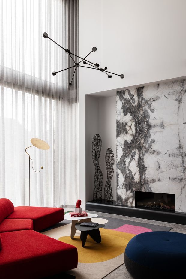 A sophisticated contemporary living room with a dramatic veined marble fireplace wall, black linear fireplace, floor-to-ceiling sheer curtains, and a sculptural black branching pendant light. Bold red sectional seating, a deep blue ottoman, colourful geometric rug, and sculptural decor add vivid accents to the neutral architectural shell.