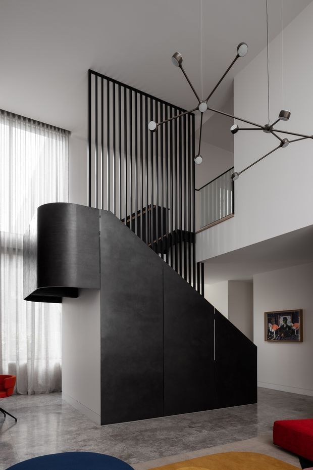 A dramatic double-height contemporary interior centered on a sculptural black staircase enclosure with vertical black balustrade slats. The space has polished grey flooring, white walls, sheer floor-to-ceiling curtains, a geometric branching pendant light, and vivid red and blue seating accents. An upper-level gallery or landing is visible beyond the stair void.