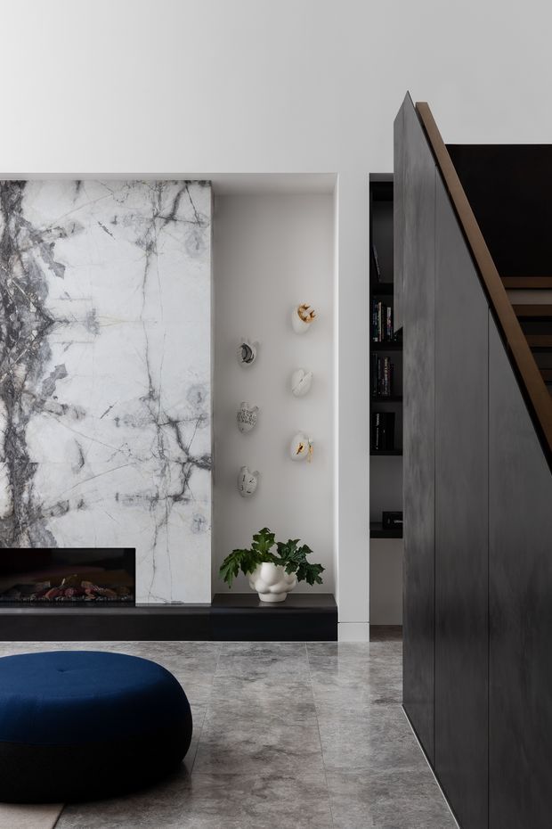 Contemporary living room interior with a dramatic veined marble feature wall and integrated fireplace, dark built-in shelving, a sculptural dark blue ottoman, and a striking charcoal staircase partition with timber-edged open treads. Grey stone flooring and neutral walls create a refined, monochromatic scheme with small decorative wall objects and a potted plant.