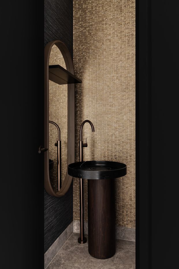 A dramatic, compact bathroom powder room featuring a freestanding dark wood cylindrical basin pedestal with a black stone or composite top, tall black curved tapware, an oval framed mirror, a small dark shelf, patterned gold-toned wall covering, dark textured wall panels, and grey stone-look floor and skirting. The space has a moody, refined, luxurious character with strong vertical detailing.