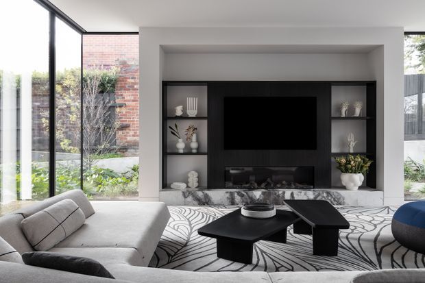 Contemporary living room with a built-in black media wall and fireplace, open shelving, pale sculptural sofas, geometric patterned rug, and black nesting coffee tables. Full-height glazing connects the room to a planted courtyard, while a brick wall is visible outside.
