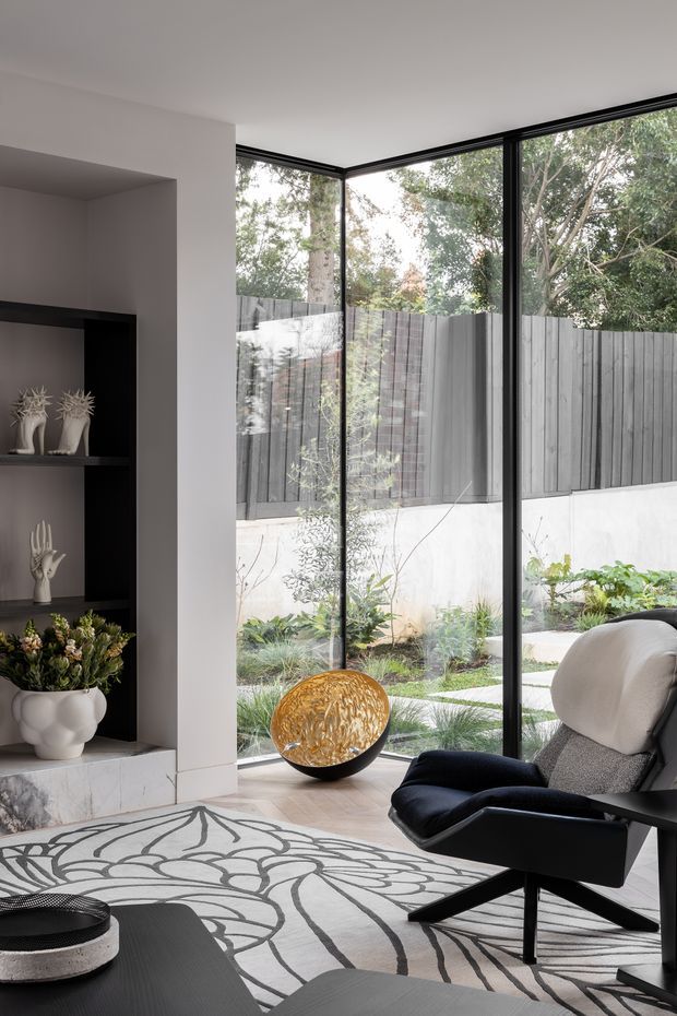 A refined contemporary living room with floor-to-ceiling black-framed corner glazing overlooking a landscaped courtyard. The room features a black built-in display niche, pale stone-clad fireplace or plinth, sculptural decor, a dark lounge chair, a patterned area rug, and a low black coffee table. The neutral interior is complemented by abundant garden views and natural light.