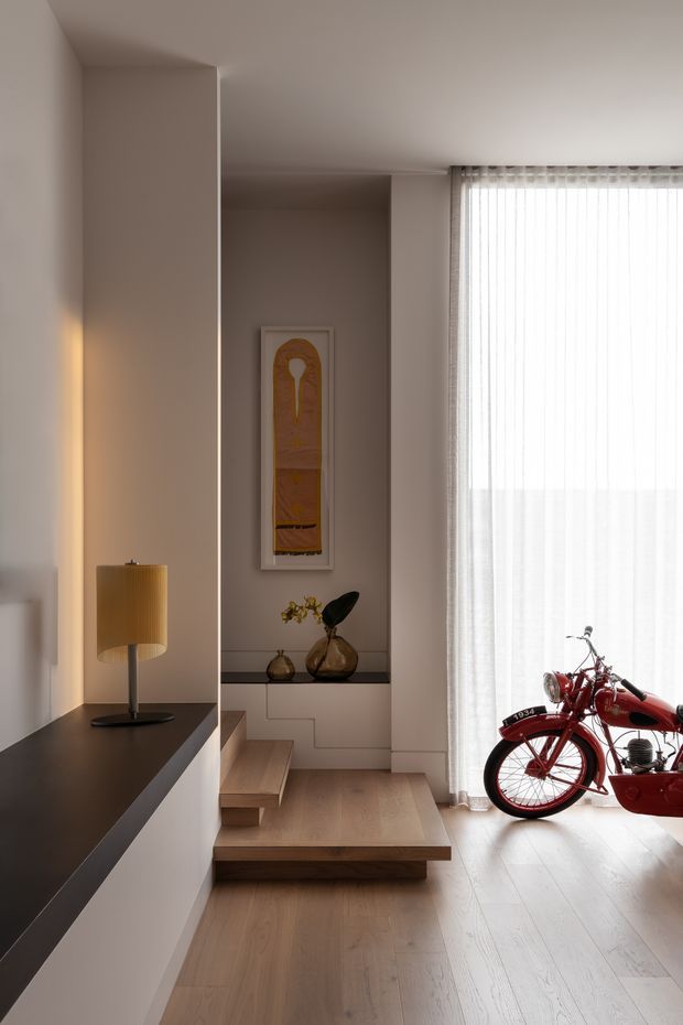 A refined minimalist interior hallway or entry zone with pale timber flooring, integrated stepped platform and stair elements, warm taupe plaster walls, and a dark built-in console with a table lamp. A large sheer-curtained window provides soft daylight, while framed artwork and decorative vessels add subtle detail. A red vintage motorcycle is displayed at the right side of the room.
