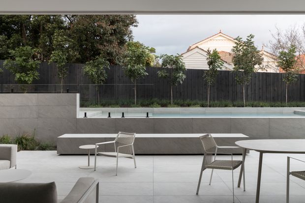 A refined contemporary outdoor courtyard or terrace viewed from beneath a covered structure. The space features pale large-format paving, a long raised rectangular pool or water feature, concrete retaining and bench walls, a dark vertical timber privacy fence, integrated planting, and minimalist outdoor dining and lounge furniture. A neighbouring traditional house is visible beyond the fence.