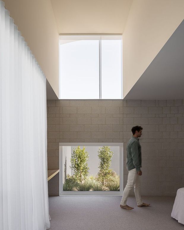 A minimalist bedroom with a dramatic high ceiling and clerestory glazing. Pale concrete block walls, a large upper window, and a low garden-facing window create a bright, sculptural interior. Sheer white curtains run along the left side, while a partially visible bed is at right and a person walks through the room. The space has a restrained, gallery-like feel with soft neutral tones and abundant natural light.