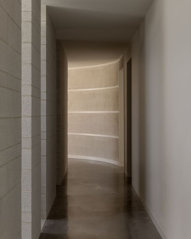 A narrow, softly lit curved corridor with smooth off-white plaster walls, a textured pale block wall on the left, and a dark polished concrete floor. The hallway bends gently around a rounded end wall, with minimal door openings and restrained architectural detailing creating a quiet, sculptural atmosphere.