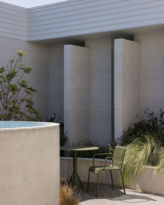 A modern courtyard or poolside terrace beside a light-toned rendered masonry building. The scene features a curved concrete pool or water feature in the foreground, pale block walls with projecting vertical fins, horizontal weatherboard-style cladding above, and a small green outdoor café table and chair surrounded by ornamental grasses and planting.