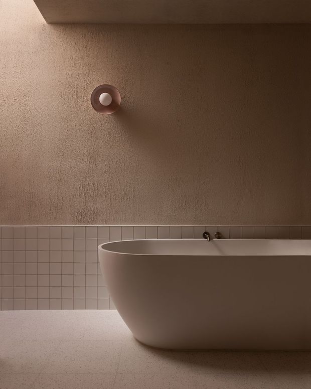 A moody, minimalist bathroom featuring a freestanding sculptural bathtub, warm taupe textured plaster walls, small square tiled wainscoting, and a lightly speckled floor. A compact circular wall light casts a soft glow and shadow above the tub.