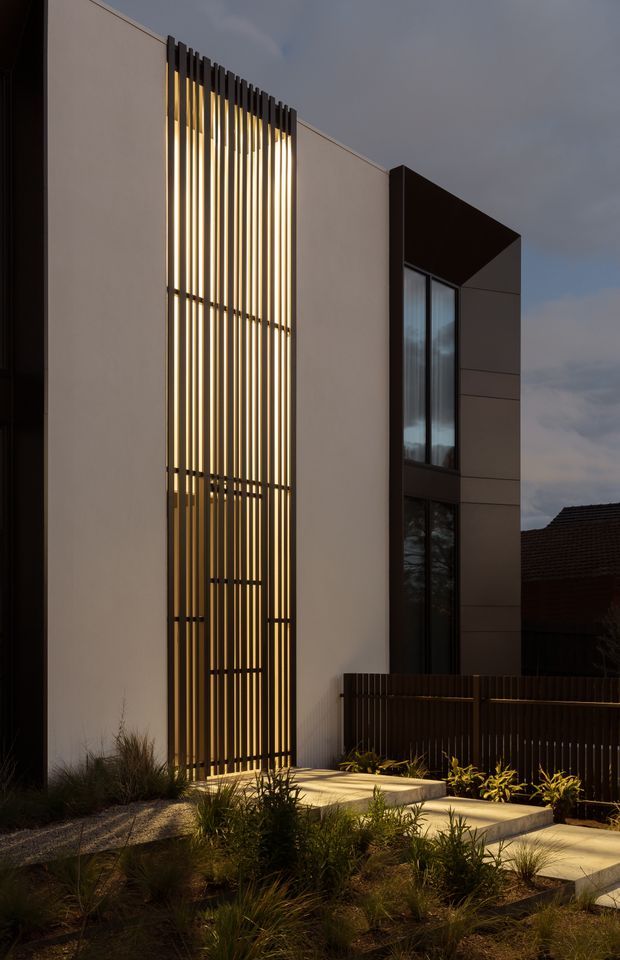 Twilight exterior photograph of a contemporary multi-storey residence. The facade combines smooth light rendered walls with dark vertical metal screening, tall narrow glazing, and a projecting dark upper volume. Warm linear lighting glows behind the slatted screen, leading to a stepped concrete entry path bordered by ornamental grasses and low planting, with a dark vertical-slat fence in the foreground.