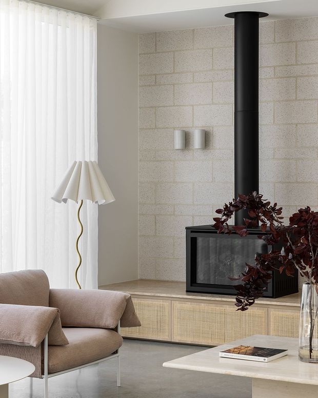 A bright, refined living room featuring a black freestanding wood-burning fireplace set against a pale exposed masonry feature wall. Full-height sheer curtains frame the window, while a sculptural floor lamp, soft beige armchair, low built-in rattan-front cabinetry, pale stone coffee table, and dark burgundy foliage create a warm contemporary composition.