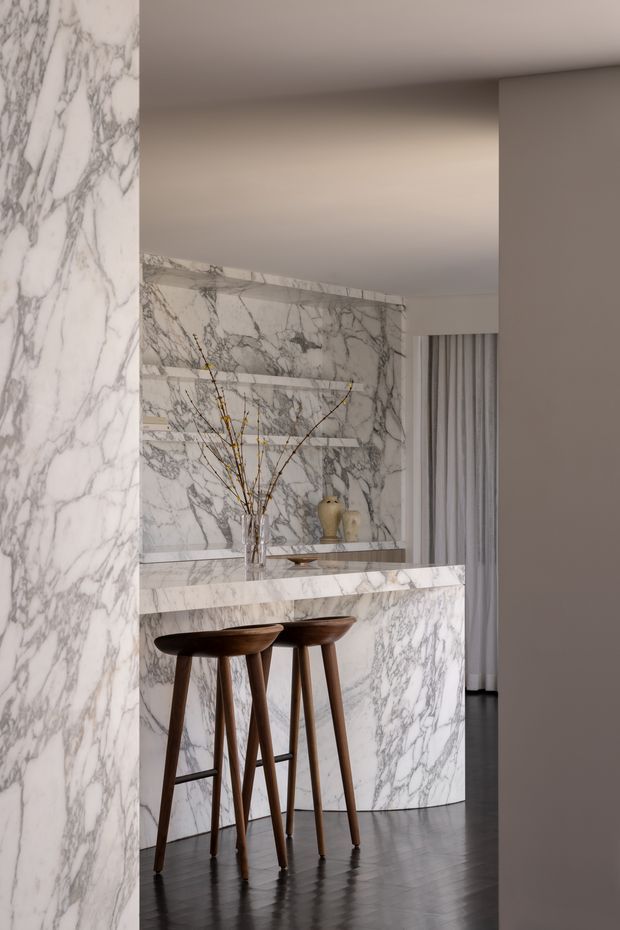 A refined kitchen or bar nook clad extensively in white veined marble, including the backsplash, shelving wall, peninsula, and surrounding wall. Two dark timber bar stools sit beneath the marble counter, with minimalist decorative vessels and branches on the shelves. The space has a warm neutral palette, dark polished flooring, soft recessed lighting, and a partially visible curtain-lined opening.