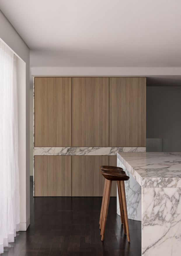 A refined contemporary kitchen with full-height warm wood cabinetry, a dramatic marble-veined island and matching backsplash, dark timber flooring, and a pair of wooden bar stools. Sheer curtains provide soft daylight along the left side, while a dark adjacent area recedes beyond the cabinetry.
