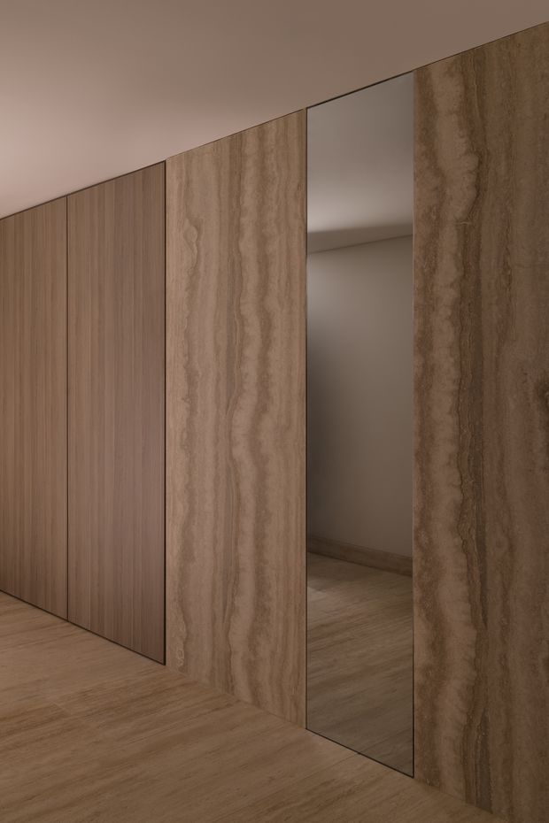 A close-up interior view of a warm, minimalist built-in wall installation. Tall vertical timber-toned cabinetry or wall panels sit beside large veined stone-look panels, with a narrow full-height mirrored opening reflecting the adjacent wall and timber floor. The space is uncluttered, monochromatic, and softly lit, with a seamless ceiling and continuous wood-look flooring.
