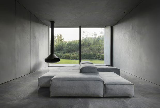 A minimalist living room with a low modular grey sectional sofa arranged around a central raised chaise or backrest. The room has monolithic grey concrete-like walls, ceiling and floor, floor-to-ceiling glazing overlooking a green landscape, and a suspended black sculptural fireplace or flue feature. The restrained palette and sparse furnishing create a calm, dramatic, gallery-like atmosphere.