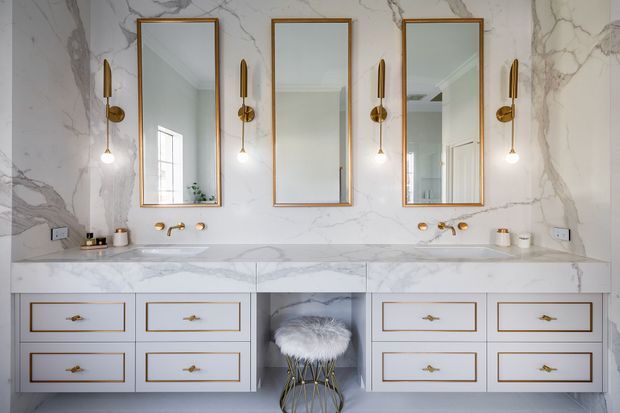 A refined double-vanity bathroom featuring a full-height white marble-look wall and benchtop, three tall brass-framed mirrors, paired brass wall sconces, twin undermount basins with brass tapware, and extensive white cabinetry with brass trim and handles. A small stool with a furry seat sits in the central open niche.