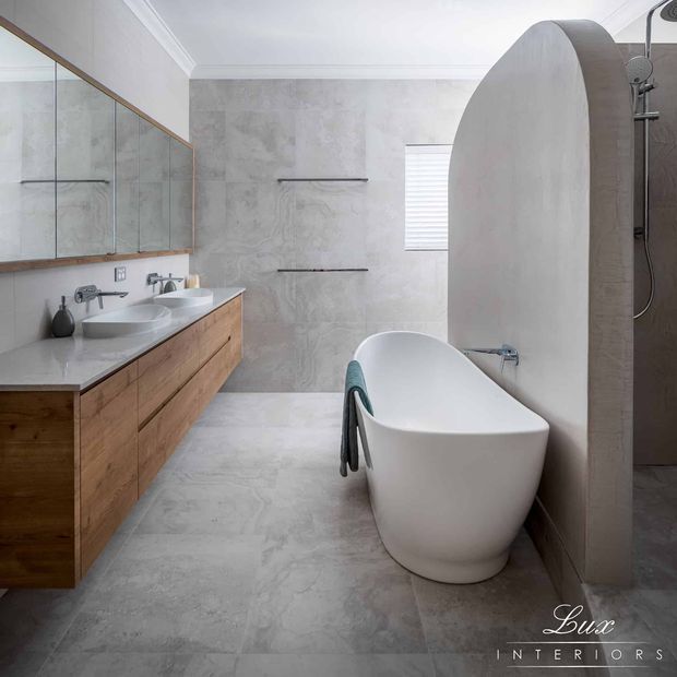 Contemporary bathroom featuring a freestanding white bathtub, double vessel sinks on a wall-mounted timber vanity, large mirrored cabinetry, pale grey stone-look wall and floor finishes, and a curved privacy partition. The space has clean lines, soft natural light, and a restrained neutral palette with warm timber accents.