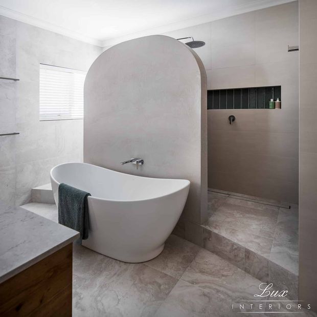 Contemporary bathroom with a freestanding white bathtub positioned beside a curved half-height partition, separating it from a walk-in shower. The room features large pale stone-look floor and wall tiles, a dark recessed shower niche, chrome fixtures, a timber vanity, and soft natural light through a shuttered window.
