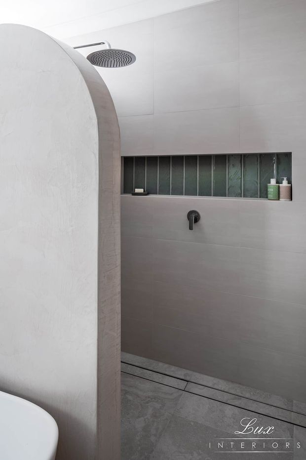Minimal contemporary walk-in shower with large pale grey wall tiles, matching stone-look floor tiles, a dark rain shower and concealed mixer, and a narrow recessed shelf with dark fluted glass backing and toiletries. A curved textured plaster privacy wall partially screens the shower.