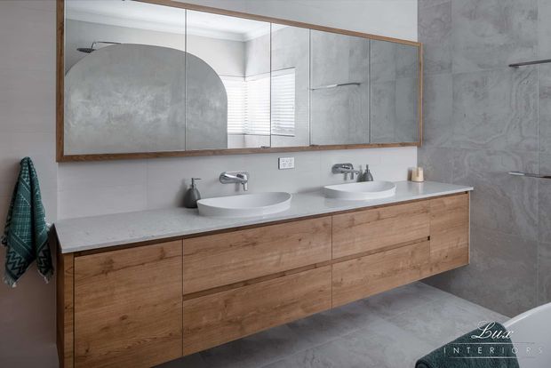 Contemporary double-vanity bathroom featuring a long floating timber-look vanity with integrated drawers, two white vessel basins, wall-mounted chrome tapware, a pale stone-look benchtop, large mirrored cabinetry with a timber frame, and large grey stone-look wall and floor tiles. A glass-enclosed shower is visible at right.