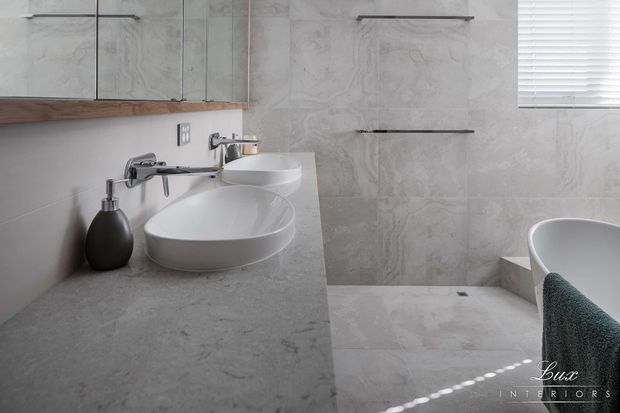 Contemporary bathroom featuring a floating grey stone vanity with a round white vessel basin, wall-mounted chrome tapware, large pale marble-look wall and floor tiles, a frameless glass shower enclosure, and a partially visible freestanding bathtub. A timber-edged mirrored cabinet and horizontal wall lighting add subtle detail.