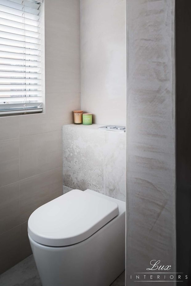 A compact contemporary bathroom featuring a modern white toilet, light grey wall tiles, a textured pale stone or concrete-look flush-panel wall, and a horizontal window with white plantation shutters. Small candles sit on the built-in ledge behind the toilet. The space has a clean, bright, neutral-toned finish.