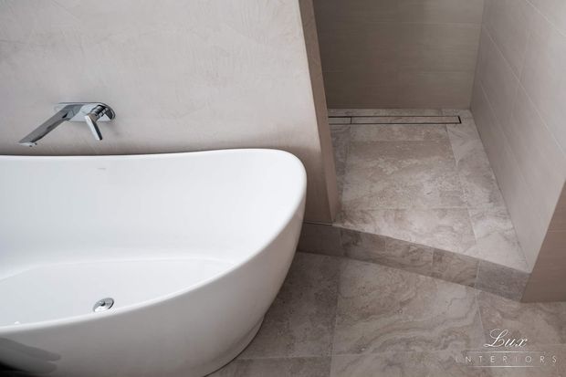 Close-up view of a refined bathroom featuring a freestanding white bathtub beside a walk-in shower. The room has pale stone-look tiled walls and flooring, a chrome wall-mounted tap, and a linear shower drain, creating a clean, neutral and understated finish.