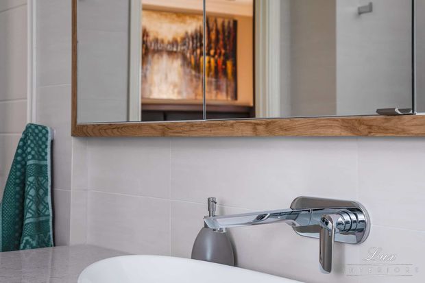 Close-up of a contemporary bathroom vanity with a white vessel basin, wall-mounted chrome tapware, white tiled wall, and a timber-framed mirrored cabinet. A patterned teal towel hangs at the side, while warm-toned artwork is reflected in the mirror.